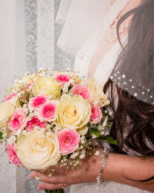 A bride holding a floral bouquet featuring large cream-colored roses and smaller pink roses, complemented by delicate white baby's breath and lush green foliage. The bouquet is arranged in a rounded s