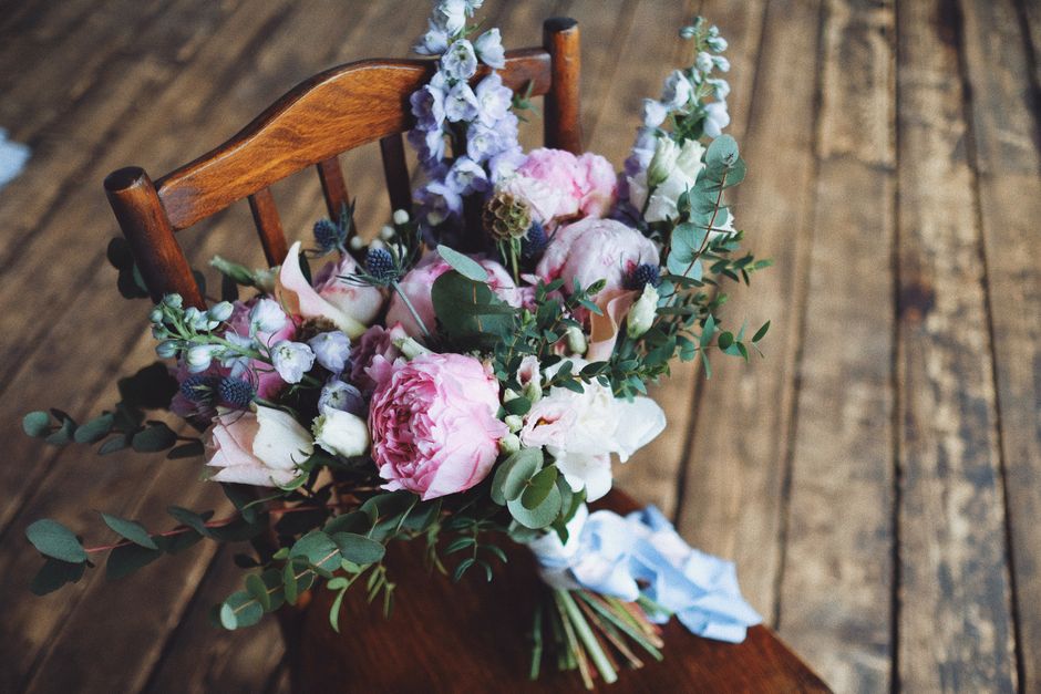 A floral arrangement placed on a wooden chair, featuring large pink peonies, white roses, and sprigs of purple thistle and eucalyptus. The bouquet is wrapped with a light blue ribbon, and the arrangem